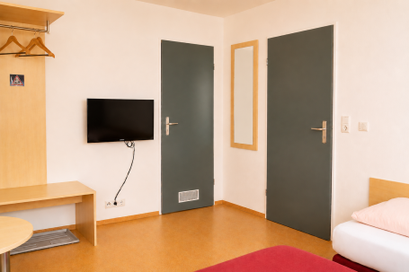 Zimmer 8 Helle, minimalistisch eingerichtete Hotelzimmer mit TV, Tisch und zwei Türen.