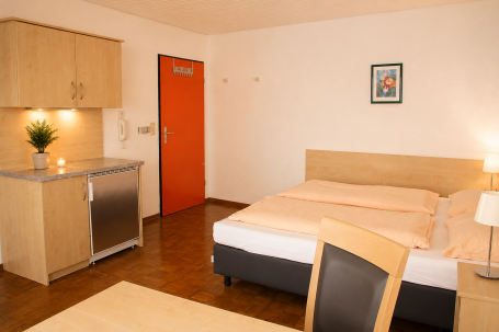 Zimmer 7 Schlafzimmer mit Doppelbett, Tisch, Stuhl, Küchenzeile und orangefarbener Tür.