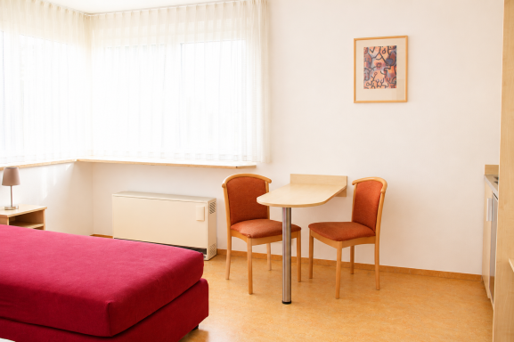 Zimmereinrichtungen Modernes Zimmer mit rotem Bett, Tisch und zwei Stühlen, Fenster mit Vorhängen.