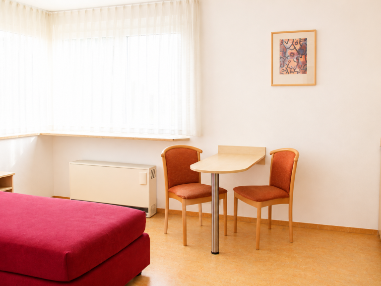 Einrichtung Gästezimmer Ein Zimmer mit einem roten Sofa, einem Tisch und zwei Stühlen vor einem Fenster.