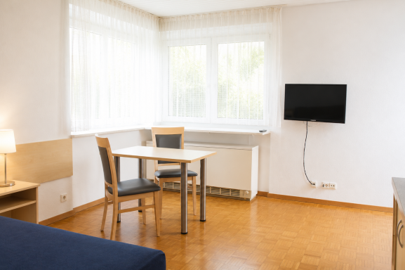 Zimmereinrichtung Heller Raum mit Tisch, Stühlen, Sofa und Wand-TV bei großen Fenstern.