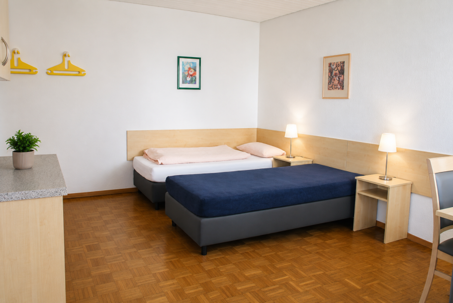 Zimmereinrichtung Helles Zimmer mit zwei Betten, Nachttischen und einem Tisch. Lampe und Pflanze vorhanden.